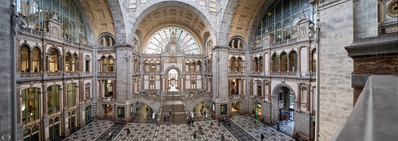 Central hall of the central station of Antwerp. A captivating panoramic image displays the train stations splendor, with an iconic arched window dominating the scene. Intricate stone and marble patterns, gray and white tile mosaic, and a birds-eye of architectural brilliance.