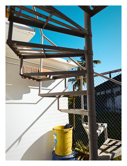 A weathered, outdoor spiral staircase made of gray metal. It ascends along a bright white wall on the left side of the frame, casting a sharp shadow on the wall. The steps of the staircase are open metal grates.

At the bottom of the staircase, near the white wall, sits a bright yellow bucket with a black plastic bag inside. The bucket rests on top of a blue container.

Behind the staircase, the scene opens up to a clear, bright blue sky. A few palm tree fronds are visible. A chain-link fence and the corner of a dark-colored building can also be seen in the background. - Google Gemini