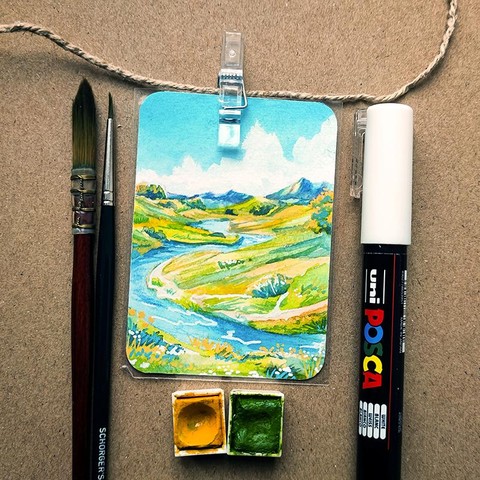 Eine kleine rechteckige Malerei, etwa so groß wie eine Visitenkarte, ist mit einer Klammer an einer Schnur befestigt und liegt auf einem braunen Hintergrund. Das Bild zeigt eine Landschaft mit sanften grünen Hügeln, einem sich schlängelnden blauen Fluss und entfernten Bergen unter einem Himmel mit weißen, fluffigen Wolken. Die Szene ist in leuchtenden Aquarellfarben gemalt. Links im Bild befinden sich zwei Pinsel, rechts ein weißer Acrylmarker. Unter dem Bild liegen zwei Aquarellnäpfchen in Gelb und Grün.
