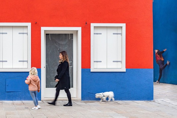 Color play with people dressed in blue and red and a white dog around a house painted in blue and red with white shutters in Burano, Italy.