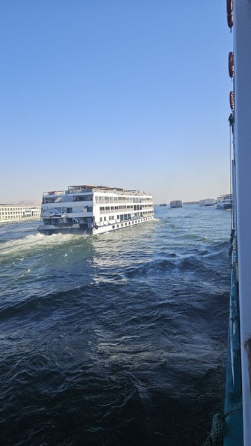 Vertical mobile phone photograph taken from the balcony of our cabin. We look along our ship to the front across the water. Right in front to our left is another cruise ship. Left of that one further ahead is another one. And straight ahead in the distance are at least five more. The riverbank of the Nile is barely visible. Everything is blocked by ships.