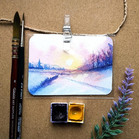 A small rectangular painting, about the size of a business card, is clipped to a string and placed on a brown background. The artwork depicts a quiet winter landscape in cool blue and violet tones, with a bright sunrise or sunset on the horizon. On the left side, there are distant buildings with a suggested church spire, while the right side shows bare trees. The foreground includes snow and a body of water reflecting the sunlight. Two paintbrushes are placed to the left, along with two watercolor pans in dark violet and yellow. A small lavender sprig lies on the right side.
