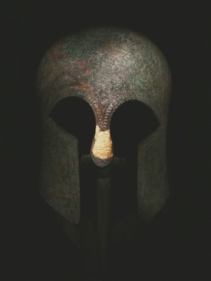 The image shows a bronze 6th century BC Corinthian helmet viewed from the front. It has cutouts for the eyes and a nose guard covered with gold sheets, which appear as a bright area. The helmet has a rounded top and a textured surface. The background is dark, and the lighting highlights the front of the helmet.
