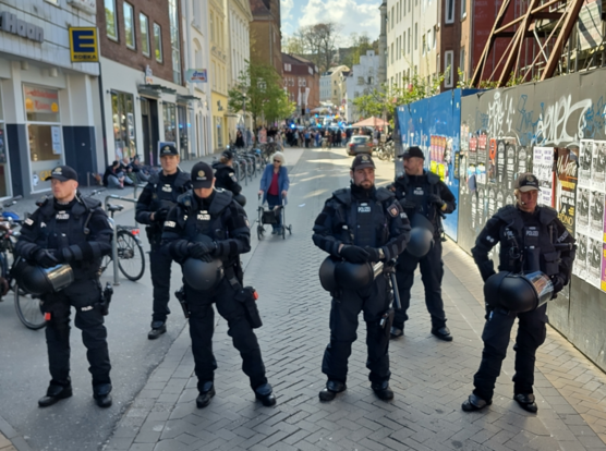 Blick die Angelburger Straße in Flensburg hinunter zum Südermarkt. Im Vordergrund eine Reihe Polizist*innen, die die Straße abriegeln. Im Hintergrund linke Demonstrant*innen, die dem rechten Demozug in der Friesischen Straße den Weg versperren, dazwischen mehrerer Polizeifahrzeuge mit Blaulicht und größere Zahl an Einsatzkräften.