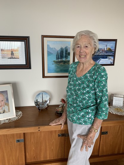 A smiling lady in a green print shirt stands in front of a sideboard with photos and knick knacks with paintings on the wall behind her including one of Moraine Lake in the Rockies.