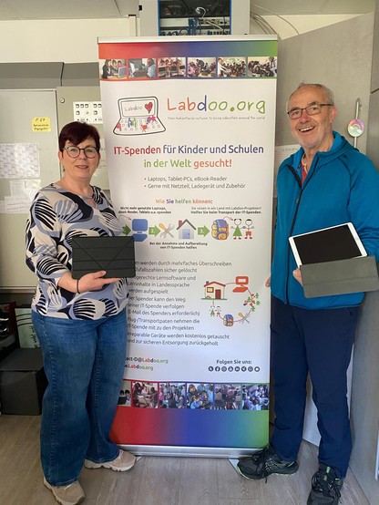 In einem Raum stehen die Leiterin des Kindergarten und ein Helfer von Labdoo vor einem Rollup von Labdoo. Beide halten jeweils ein Tablet in den Händen und schauen in die Kamera.