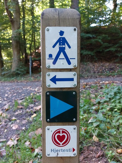 Ein Wegweiser für einen Wanderweg entlang der Küste in Dänemark. Auf dem Holzpfosten sind vier Schilder angebracht. Die vier Piktogramme von oben nach unten: Auf weißem Grund ein dunkelblauer Mann in einer Zöllner-Uniform, ein blauer Pfeil, der nach links zeigt, ein nach rechts weisendes türkises Dreieck auf schwarzem Grund, und auf Weiß ein rotes Herz in einem roten Kreis und das Wort Hjertesti.
🇬🇧 A signpost for a hiking trail along the coast in Denmark. There are four signs on the wooden post. The four pictograms from top to bottom: On a white background a dark blue man in a customs officer's uniform, a blue arrow pointing to the left, a turquoise triangle pointing to the right on a black background, and on white a red heart in a red circle and the word Hjertesti.