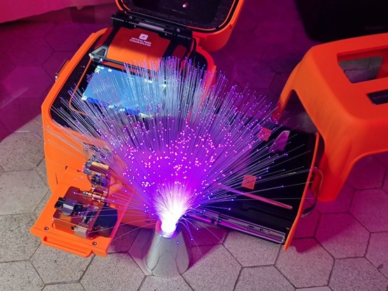 Im Vordergrund viele aufgefächerte Glasfasern die mit Lila-Licht beleuchtet sind. Im Hintergrund ein Spleißgerät für Glasfasern.