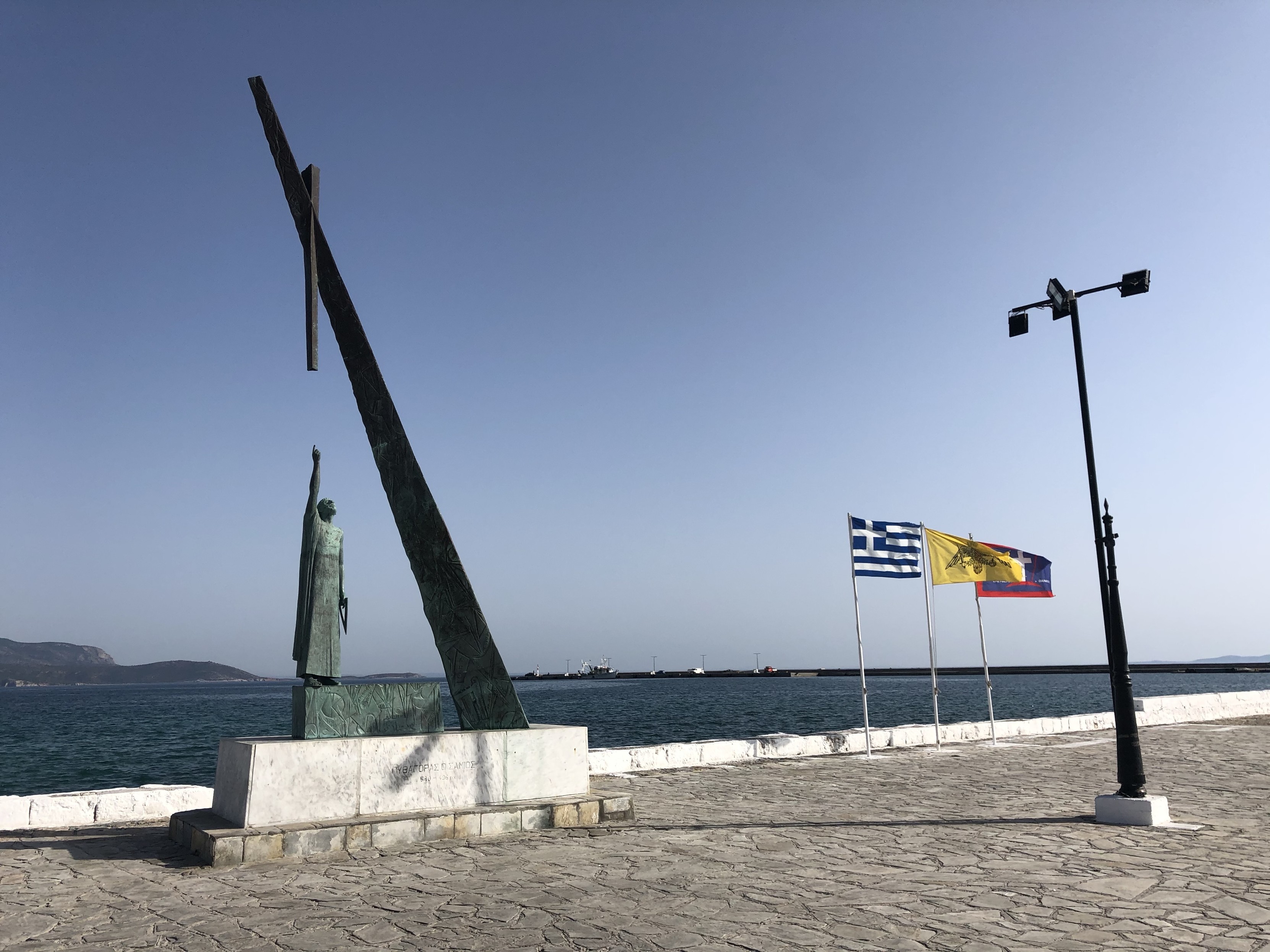 Statue von Pythagoras (ja, der mit dem rechtwinkligen Dreieck) in Pythagorio, Samos, Griechenland. Die Bronzestatue, die einen erhobenen Arm hält, steht auf einer Strandpromenade. Im Hintergrund werden drei Flaggen angezeigt: eine griechische Flagge, eine gelbe Flagge mit einem Wappen und eine rote Flagge. Im Hintergrund wolkenloser Himmel und das Mittelmeer. Statue von Pythagoras (ja, der mit dem rechtwinkligen Dreieck) in Pythagorio, Samos, Griechenland. Die Bronzestatue, die einen erhobenen Arm hält, steht auf einer Strandpromenade. Im Hintergrund werden drei Flaggen angezeigt: eine griechische Flagge, eine gelbe Flagge mit einem Wappen und eine rote Flagge. Im Hintergrund wolkenloser Himmel und das Mittelmeer.