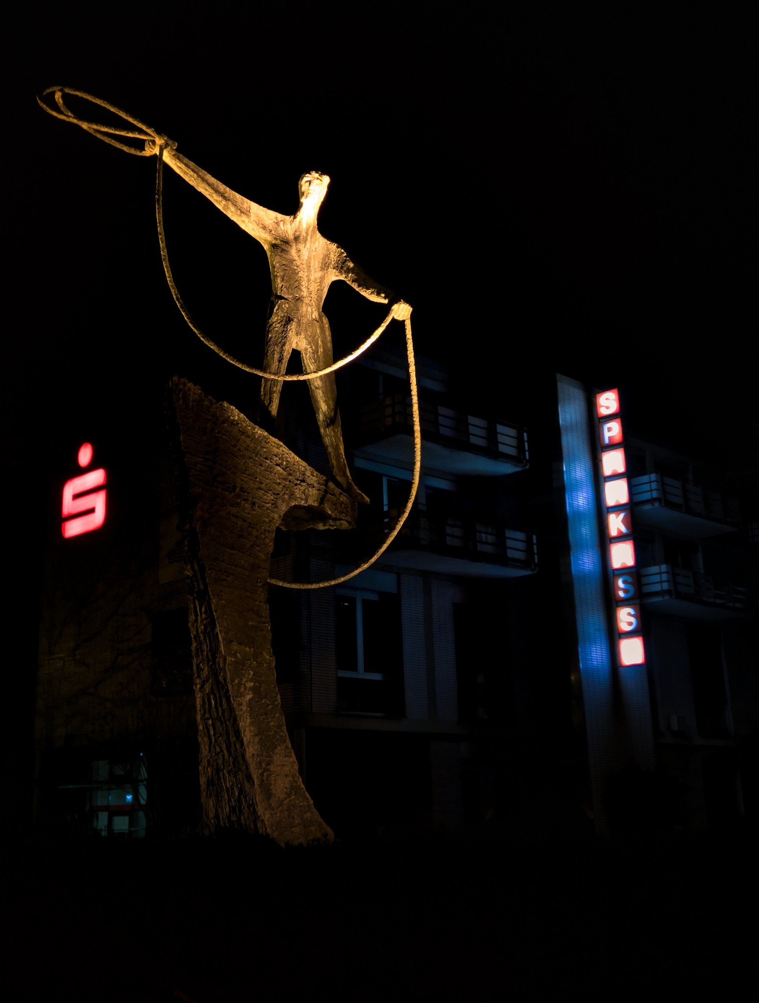Nächtliches Foto von Robert Metallstatue, die in Wildwest-Manier ein Lasso schwingt. Im Hintergrund sind die Leuchtzeichen einer Sparkasse zu sehen. Nächtliches Foto von Robert Metallstatue, die in Wildwest-Manier ein Lasso schwingt. Im Hintergrund sind die Leuchtzeichen einer Sparkasse zu sehen.