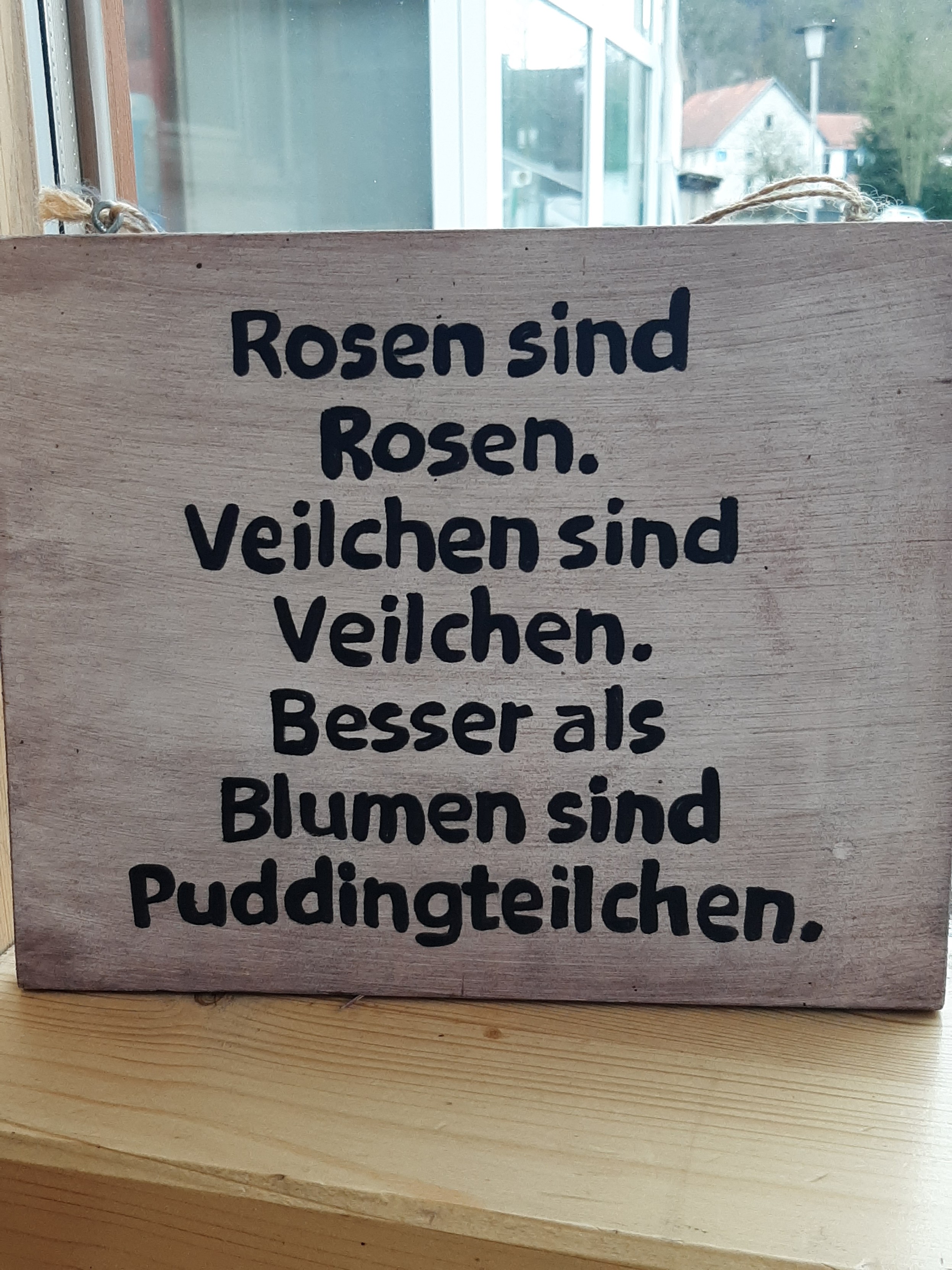 Holzschild mit der Aufschrift:
"Rosen sind Rosen.
Veilchen sind Veilchen.
Besser als Blumen sind
Pudding Teilchen."