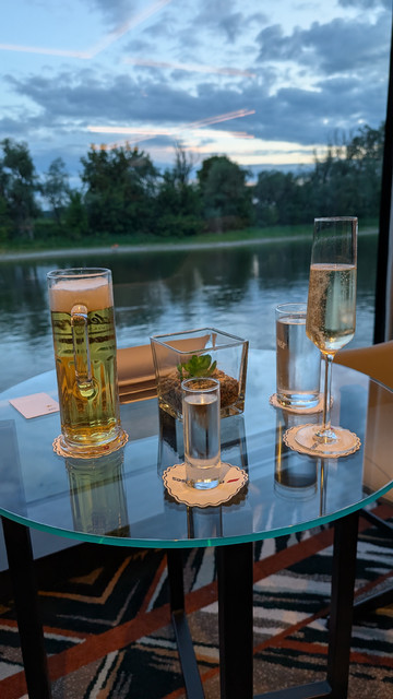 Auf einem runden Glastisch in der Lounge des Schiffes stehen vier Gläser mit verschiedenen Getränken. Im Hintergrund das Wasser der Donau und grün bewachsenes Ufer. 