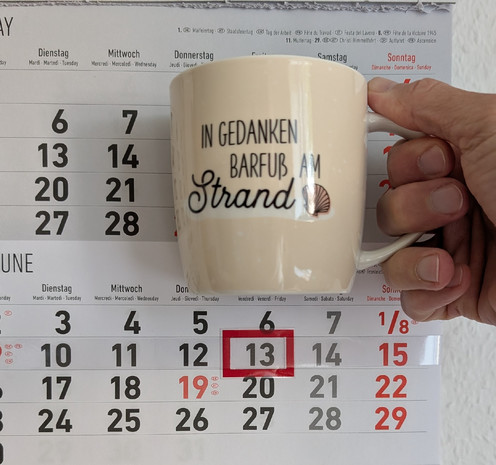 Meine rechte Hand hält einen Porzellan-Kaffeebecher. Seine Farbe ist beige. In schwarzer Schrift steht auf ihm: In Gedanken barfuß am Strand, gefolgt von einer Zeichnung einer bräunlichen Meermuschel. Im Hintergrund hängt ein 3-Monats-Wandkalender an der Wand mit dem rot umrandeten heutigen Tag. 
