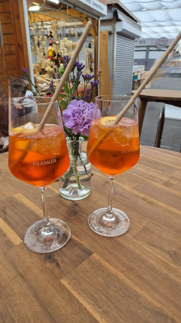 Zwei Gläser Aperol Spritz auf einem Holztisch. Dahinter ein kleines, lilafarbenes Blumengesteck in einer Glasvase. 