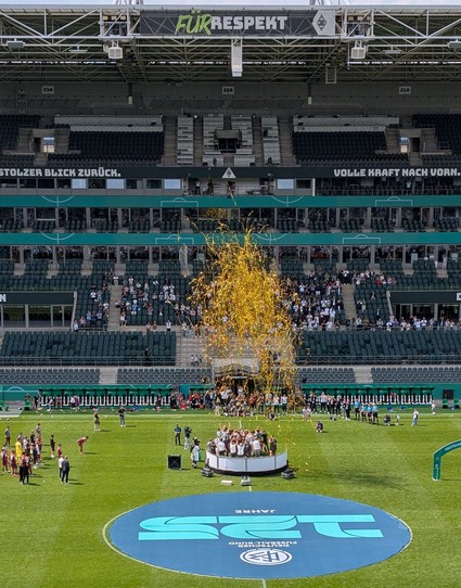Kamera ausgelöst Bruchteile nach Abschuss der Goldregen-Kanonen. Die goldfarbenen Schnipsel schweben zentral über den auf einem Podest feiernden U17-Spielern von Borussia Mönchengladbach. Vor dem Podest liegt im Mittelkreis des Spielfeldes ein rundes Banner zum 125 jährigen Bestehen des DFBs. Im Hintergrund die Haupttribüne mit einigen Zuschauenden sowie einem zentral darüber hängenden rechteckigen Banner mit der Aufschrift "Für Respekt" unter dem Stadiondach.