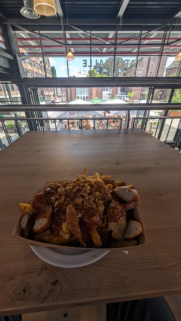 Im Vordergrund auf einen quadratischen Holztisch eine Schale mit Currywurst auf Pommes. Da ich bei der Aufnahme auf der offenen Empore sitze, sind im Hintergrund die Tische mit Gästen im Außenbereich vor der Markthalle in Mönchengladbach. 