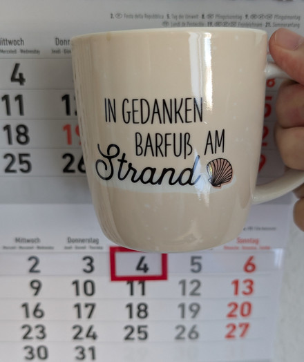 Meine rechte Hand hält einen Porzellan-Kaffeebecher. Seine Farbe ist beige. In schwarzer Schrift steht auf ihm: In Gedanken barfuß am Strand, gefolgt von einer Zeichnung einer bräunlichen Meermuschel. Im Hintergrund hängt ein 3-Monats-Wandkalender an der Wand mit dem rot umrandeten heutigen Tag. 