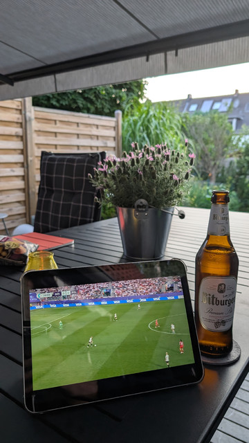 Das Spiel Deutschland gegen Polen läuft auf einem Tablet. Neben dem Gerät steht eine Flasche Pils. Dahinter auf dem Terrassentisch eine Schale mit der Pflanze. Im Hintergrund der Garten. 