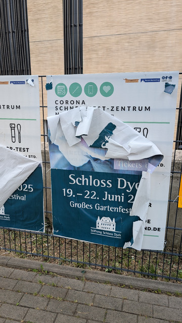 Ein Papier-Plakat an einem Zaun vor einem Supermarkt, das für Festspiele im Juni 2025 wirbt, blättert von seinem Plastik-Trägermaterial ab. Zum Vorschein kommen dadurch die Hinweise auf Corona Schnelltest-Stationen in Neuss. 
