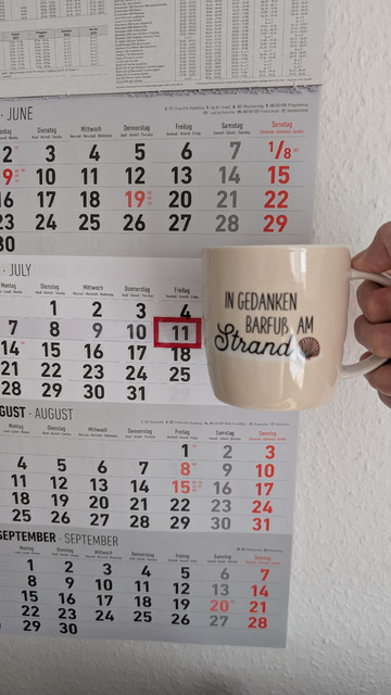 Meine rechte Hand hält einen Porzellan-Kaffeebecher. Seine Farbe ist beige. In schwarzer Schrift steht auf ihm: In Gedanken barfuß am Strand, gefolgt von einer Zeichnung einer bräunlichen Meermuschel. Im Hintergrund hängt ein 3-Monats-Wandkalender an der Wand mit dem rot umrandeten heutigen Tag. 