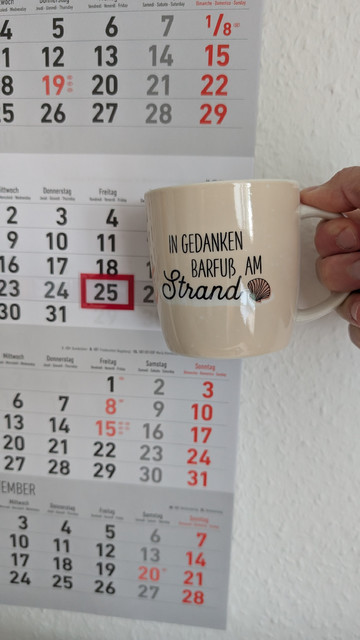 Meine rechte Hand hält einen Porzellan-Kaffeebecher. Seine Farbe ist beige. In schwarzer Schrift steht auf ihm: In Gedanken barfuß am Strand, gefolgt von einer Zeichnung einer bräunlichen Meermuschel. Im Hintergrund hängt ein 3-Monats-Wandkalender an der Wand mit dem rot umrandeten heutigen Tag. 