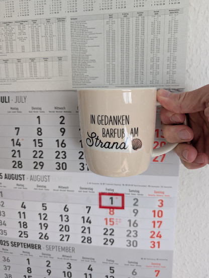 Meine rechte Hand hält einen Porzellan-Kaffeebecher. Seine Farbe ist beige. In schwarzer Schrift steht auf ihm: In Gedanken barfuß am Strand, gefolgt von einer Zeichnung einer bräunlichen Meermuschel. Im Hintergrund hängt ein 3-Monats-Wandkalender an der Wand mit dem rot umrandeten heutigen Tag. 