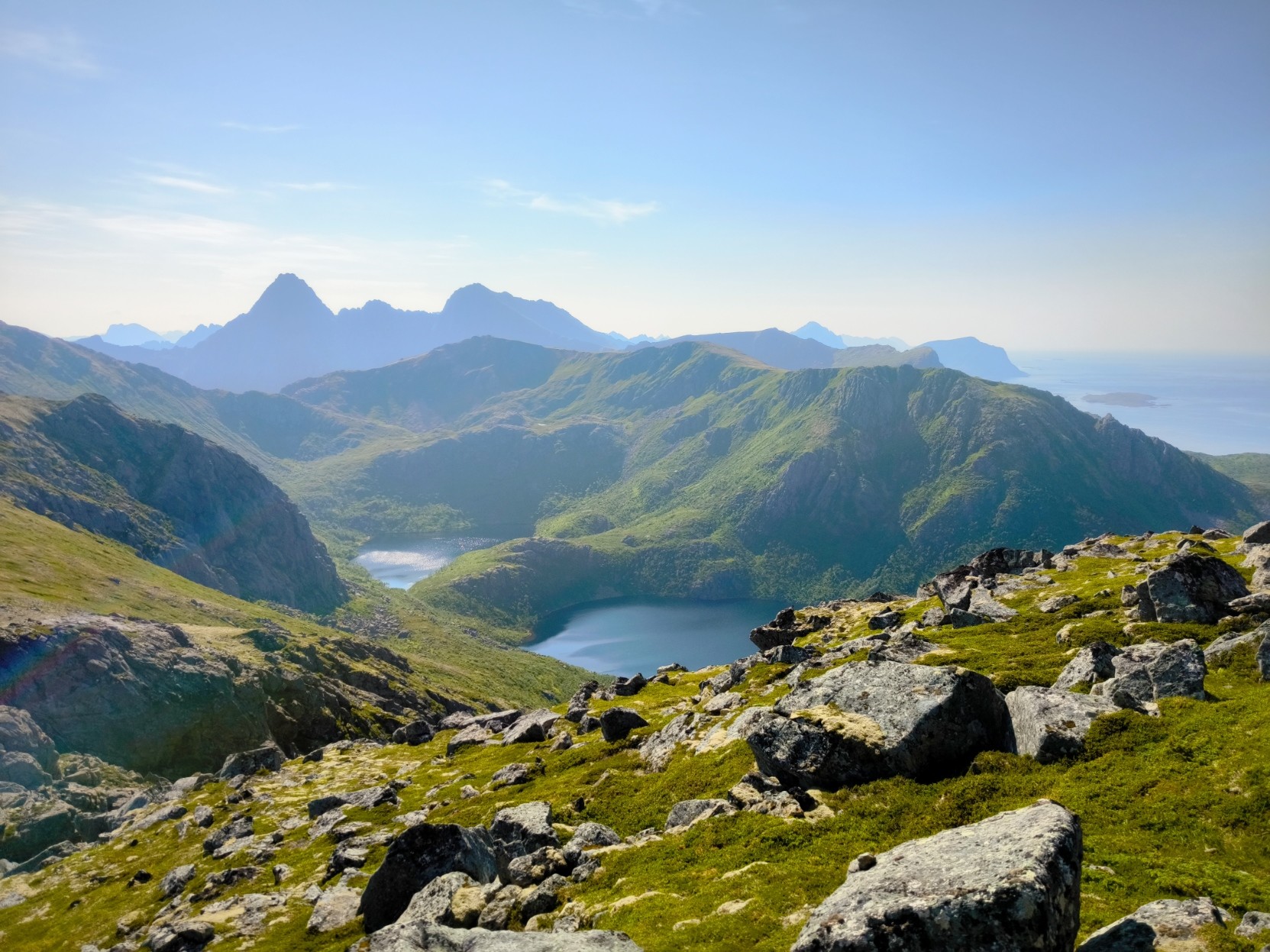 The image depicts a breathtaking mountainous landscape under a clear blue sky. In the foreground, there is a rocky terrain with patches of green grass and moss, scattered with large boulders. The midground features a serene lake nestled between rolling hills, reflecting the surrounding greenery and sky. The background showcases a range of mountains with varying elevations, some with sharp peaks and others with more rounded forms. The mountains are covered in lush green vegetation, indicating a healthy, natural environment. The distant horizon reveals a body of water, possibly an ocean or a large sea, with a faint outline of landmasses. The overall scene is tranquil and picturesque, with the interplay of light and shadow enhancing the natural beauty of the landscape.