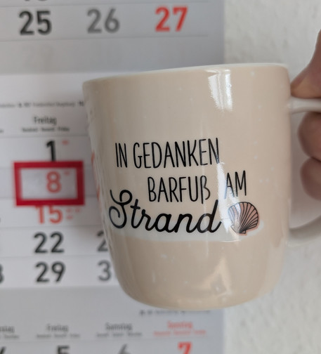 Meine rechte Hand hält einen Porzellan-Kaffeebecher. Seine Farbe ist beige. In schwarzer Schrift steht auf ihm: In Gedanken barfuß am Strand, gefolgt von einer Zeichnung einer bräunlichen Meermuschel. Im Hintergrund hängt ein 3-Monats-Wandkalender an der Wand mit dem rot umrandeten heutigen Tag. 