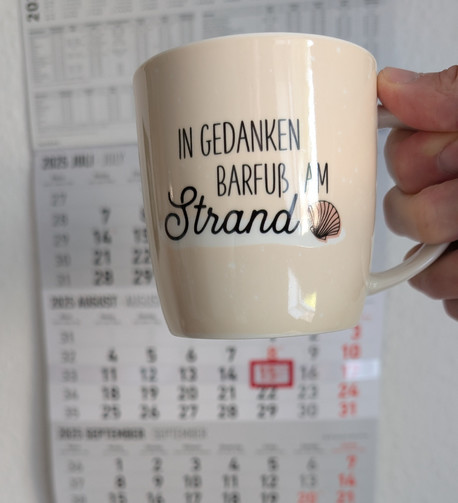 Meine rechte Hand hält einen Porzellan-Kaffeebecher. Seine Farbe ist beige. In schwarzer Schrift steht auf ihm: In Gedanken barfuß am Strand, gefolgt von einer Zeichnung einer bräunlichen Meermuschel. Im Hintergrund hängt ein 3-Monats-Wandkalender an der Wand mit dem rot umrandeten heutigen Tag. 