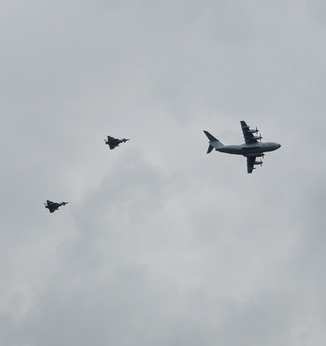 Ein viermotoriges Militärflugzeug, eventuell eine Hercules, gefolgt von zwei Begleit-Kampfjets, vor grauem Himmel über Kappeln. 