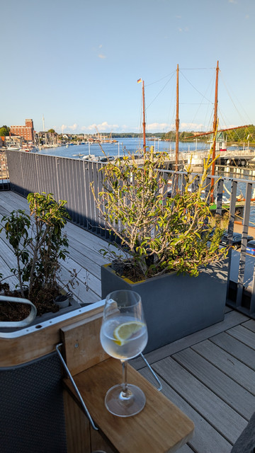 Der Ausblick auf die Schlei und den Ort Kappeln aus dem Strandkorb heraus. Im Vordergrund steht auf dem Klapptischchen des Strandkorbs ein Weinglas, das mit einem klaren Getränk mit halber Zitronenscheibe gefüllt ist. 