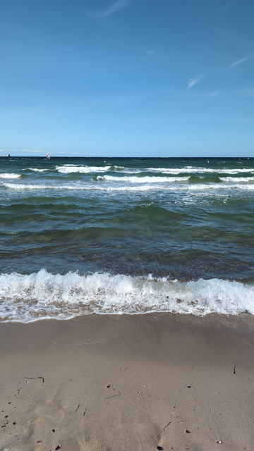 Blick vom Strand auf die Ostsee. Wellen mit weißen Schaumkronen rollen an den Sand. Der Himmel ist blau und fast wolkenlos. 