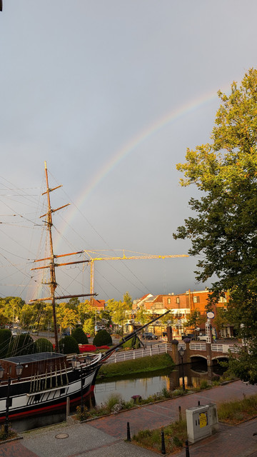 Im Vordergrund ein altes Segelschiff, das im Kanal vor dem Hotel festgemacht ist. Dahinter grauer Himmel, in dem sich ein Regenbogen abzeichnet. 