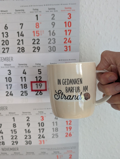 Meine rechte Hand hält einen Porzellan-Kaffeebecher. Seine Farbe ist beige. In schwarzer Schrift steht auf ihm: In Gedanken barfuß am Strand, gefolgt von einer Zeichnung einer bräunlichen Meermuschel. Im Hintergrund hängt ein 3-Monats-Wandkalender an der Wand mit dem rot umrandeten heutigen Tag. 
