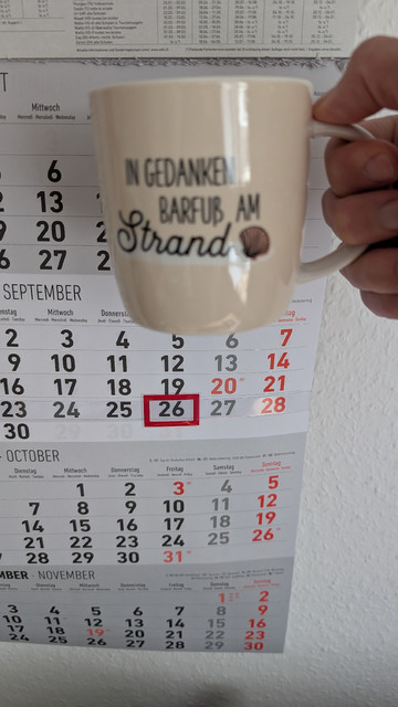 Meine rechte Hand hält einen Porzellan-Kaffeebecher. Seine Farbe ist beige. In schwarzer Schrift steht auf ihm: In Gedanken barfuß am Strand, gefolgt von einer Zeichnung einer bräunlichen Meermuschel. Im Hintergrund hängt ein 3-Monats-Wandkalender an der Wand mit dem rot umrandeten heutigen Tag. 