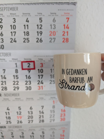 Meine rechte Hand hält einen Porzellan-Kaffeebecher. Seine Farbe ist beige. In schwarzer Schrift steht auf ihm: In Gedanken barfuß am Strand, gefolgt von einer Zeichnung einer bräunlichen Meermuschel. Im Hintergrund hängt ein 3-Monats-Wandkalender an der Wand mit dem rot umrandeten heutigen Tag. 