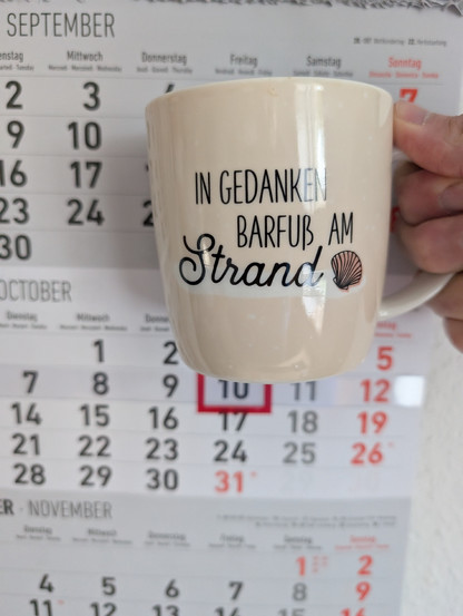Meine rechte Hand hält einen Porzellan-Kaffeebecher. Seine Farbe ist beige. In schwarzer Schrift steht auf ihm: In Gedanken barfuß am Strand, gefolgt von einer Zeichnung einer bräunlichen Meermuschel. Im Hintergrund hängt ein 3-Monats-Wandkalender an der Wand mit dem rot umrandeten heutigen Tag. 