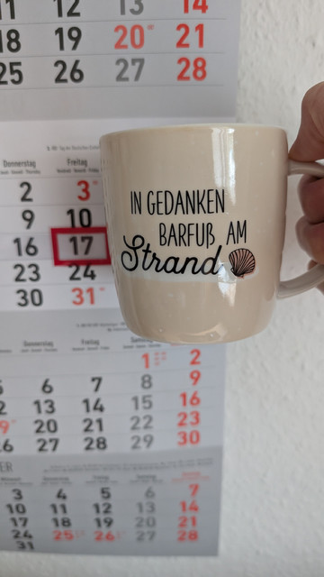 Meine rechte Hand hält einen Porzellan-Kaffeebecher. Seine Farbe ist beige. In schwarzer Schrift steht auf ihm: In Gedanken barfuß am Strand, gefolgt von einer Zeichnung einer bräunlichen Meermuschel. Im Hintergrund hängt ein 3-Monats-Wandkalender an der Wand mit dem rot umrandeten heutigen Tag. 