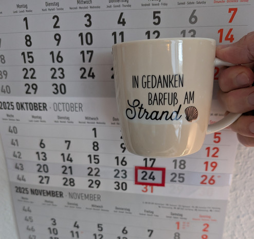 Meine rechte Hand hält einen Porzellan-Kaffeebecher. Seine Farbe ist beige. In schwarzer Schrift steht auf ihm: In Gedanken barfuß am Strand, gefolgt von einer Zeichnung einer bräunlichen Meermuschel. Im Hintergrund hängt ein 3-Monats-Wandkalender an der Wand mit dem rot umrandeten heutigen Tag. 