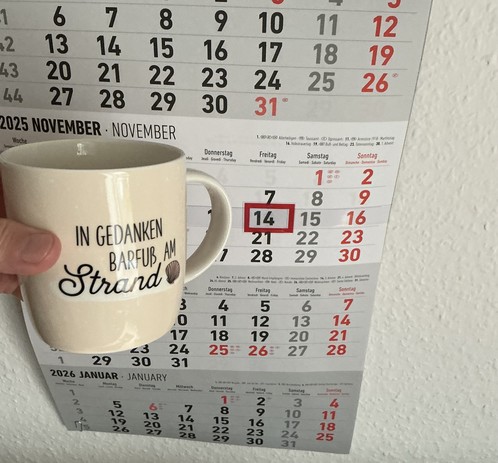 Meine rechte Hand hält einen Porzellan-Kaffeebecher. Seine Farbe ist beige. In schwarzer Schrift steht auf ihm: In Gedanken barfuß am Strand, gefolgt von einer Zeichnung einer bräunlichen Meermuschel. Im Hintergrund hängt ein 3-Monats-Wandkalender an der Wand mit dem rot umrandeten heutigen Tag. 