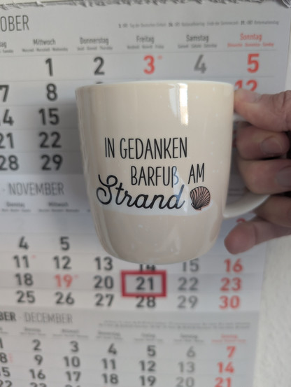 Meine rechte Hand hält einen Porzellan-Kaffeebecher. Seine Farbe ist beige. In schwarzer Schrift steht auf ihm: In Gedanken barfuß am Strand, gefolgt von einer Zeichnung einer bräunlichen Meermuschel. Im Hintergrund hängt ein 3-Monats-Wandkalender an der Wand mit dem rot umrandeten heutigen Tag. 