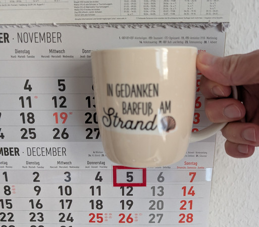 Meine rechte Hand hält einen Porzellan-Kaffeebecher. Seine Farbe ist beige. In schwarzer Schrift steht auf ihm: In Gedanken barfuß am Strand, gefolgt von einer Zeichnung einer bräunlichen Meermuschel. Im Hintergrund hängt ein 3-Monats-Wandkalender an der Wand mit dem rot umrandeten heutigen Tag. 