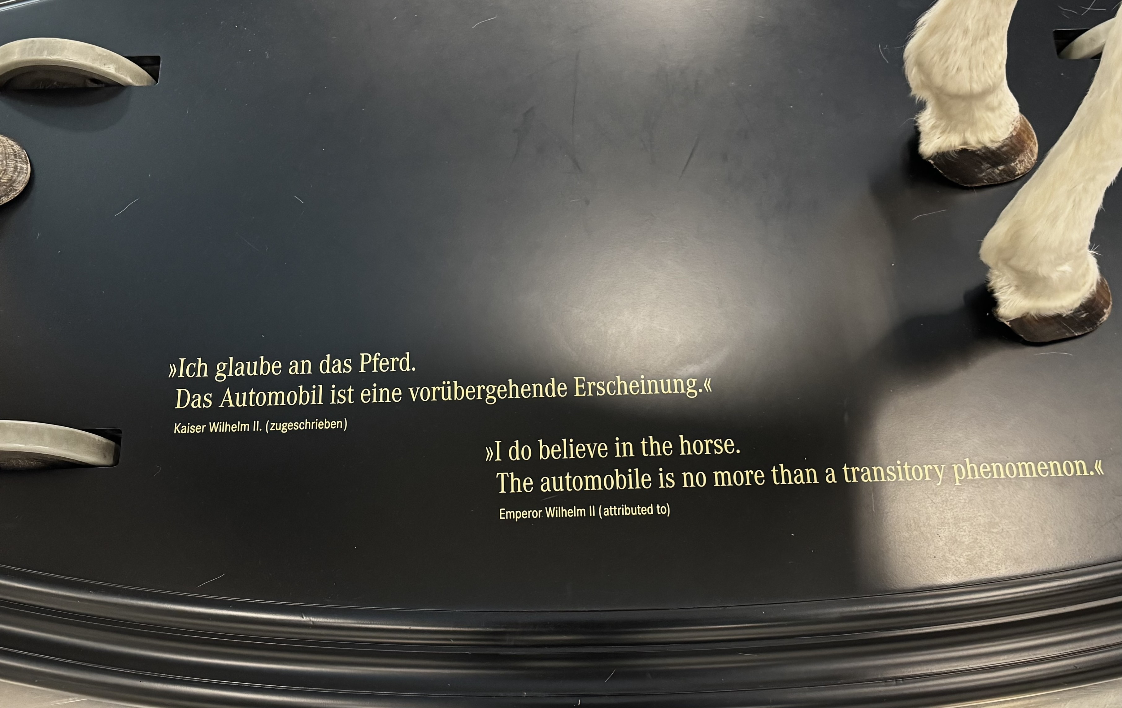 Das Bild zeigt ein dekoratives Display mit dem Zitat "Ich glaube an das Pferd. Das Automobil ist nicht mehr als ein vorübergehendes Phänomen." Kaiser Wilhelm II. zugeschrieben. Die Inschrift ist sowohl auf Deutsch als auch auf Englisch geschrieben und zeigt einen schwarzen Hintergrund. Auf den Seiten des Bildes sind noch die Hufe des weissen Pferdes zu erkennen.