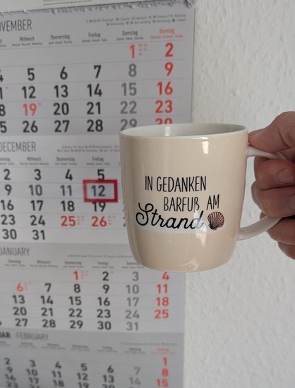 Meine rechte Hand hält einen Porzellan-Kaffeebecher. Seine Farbe ist beige. In schwarzer Schrift steht auf ihm: In Gedanken barfuß am Strand, gefolgt von einer Zeichnung einer bräunlichen Meermuschel. Im Hintergrund hängt ein 3-Monats-Wandkalender an der Wand mit dem rot umrandeten heutigen Tag. 
