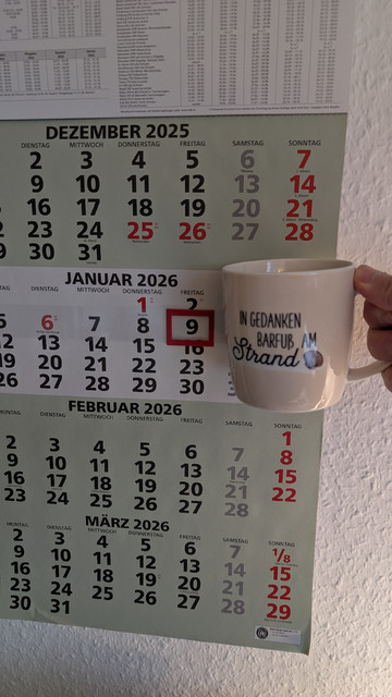 Meine rechte Hand hält einen Porzellan-Kaffeebecher. Seine Farbe ist beige. In schwarzer Schrift steht auf ihm: In Gedanken barfuß am Strand, gefolgt von einer Zeichnung einer bräunlichen Meermuschel. Im Hintergrund hängt ein 3-Monats-Wandkalender an der Wand mit dem rot umrandeten heutigen Tag. 
