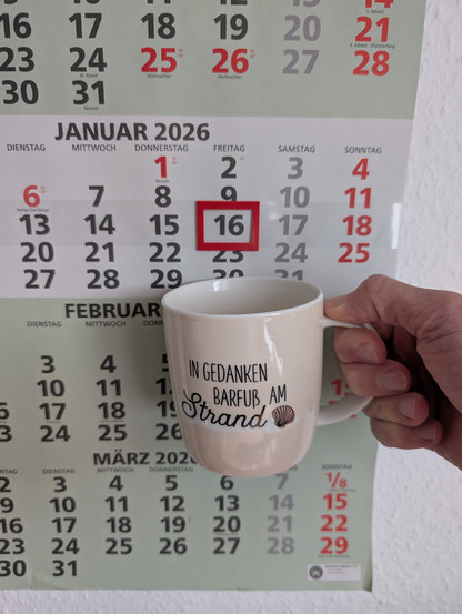Meine rechte Hand hält einen Porzellan-Kaffeebecher. Seine Farbe ist beige. In schwarzer Schrift steht auf ihm: In Gedanken barfuß am Strand, gefolgt von einer Zeichnung einer bräunlichen Meermuschel. Im Hintergrund hängt ein 3-Monats-Wandkalender an der Wand mit dem rot umrandeten heutigen Tag. 