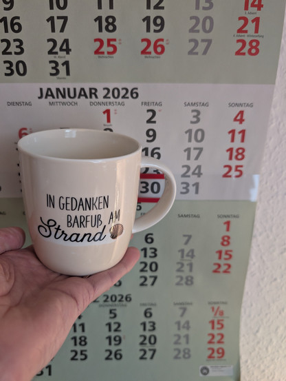 Auf den Fingern meiner linken Hand steht ein Porzellan-Kaffeebecher. Seine Farbe ist beige. In schwarzer Schrift steht auf ihm: In Gedanken barfuß am Strand, gefolgt von einer Zeichnung einer bräunlichen Meermuschel. Im Hintergrund hängt ein 3-Monats-Wandkalender an der Wand mit dem rot umrandeten heutigen Tag.  Er befindet sich direkt hinter - also eigentlich im - Henkel der Tasse. 