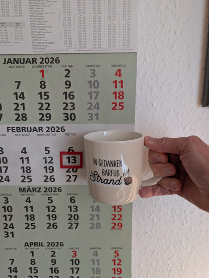 Meine rechte Hand hält einen Porzellan-Kaffeebecher. Seine Farbe ist beige. In schwarzer Schrift steht auf ihm: In Gedanken barfuß am Strand, gefolgt von einer Zeichnung einer bräunlichen Meermuschel. Im Hintergrund hängt ein 3-Monats-Wandkalender an der Wand mit dem rot umrandeten heutigen Tag. 