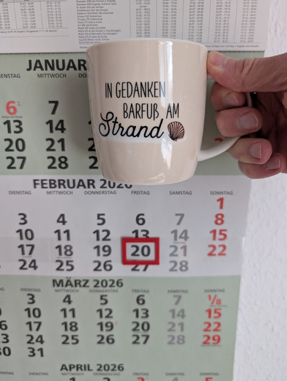 Meine rechte Hand hält einen Porzellan-Kaffeebecher. Seine Farbe ist beige. In schwarzer Schrift steht auf ihm: In Gedanken barfuß am Strand, gefolgt von einer Zeichnung einer bräunlichen Meermuschel. Im Hintergrund hängt ein 3-Monats-Wandkalender an der Wand mit dem rot umrandeten heutigen Tag. 