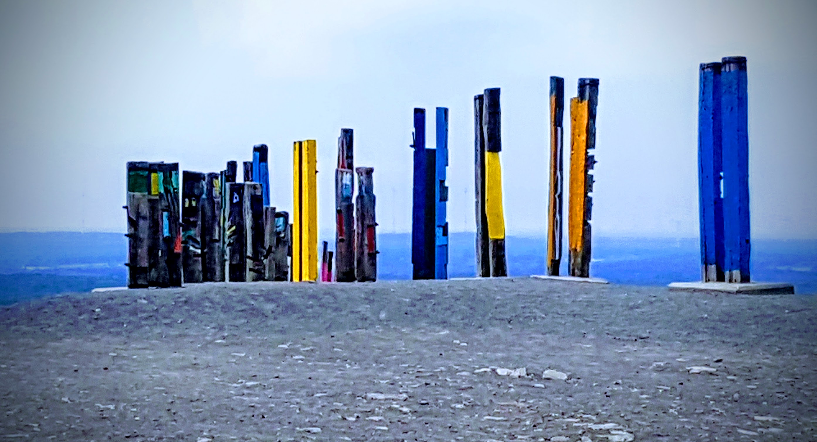 Kunstinstallation „Totems“ auf der Halde Haniel bei Bottrop/Oberhausen: Eine Reihe farbig bemalter, senkrecht stehender Holzstelen aus alten Bahnschwellen in kräftigen Tönen wie Gelb, Blau, Grün und Schwarz erhebt sich auf dem flachen Gipfelgelände einer ehemaligen Kohlehalde. Die Skulpturen des baskischen Künstlers Agustín Ibarrola stehen hier seit 2002 als öffentliches, dauerhaftes Kunstwerk und verbinden die industrielle Bergbaugeschichte des Ruhrgebiets mit Natur und Abstraktion. Die farbenfrohen Totems wirken als markantes Landmark und setzen lebendige Akzente vor dem weiten, hellen Himmel.